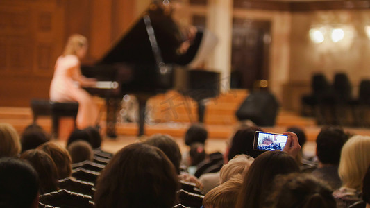 钢琴演奏节目单摄影照片_音乐厅的观众在表演钢琴女孩时 — 人们在智能手机上拍摄表演