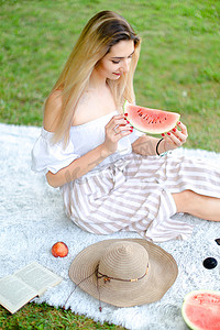 年轻的白人女孩吃西瓜，坐在格子上，靠近水果和帽子，背景是草。