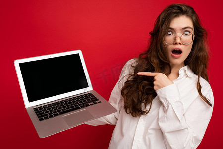 震惊的美丽深色卷发年轻女性说哇，拿着电脑笔记本电脑，戴着眼镜白衬衫，看着红墙背景中突显的相机
