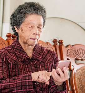 亚洲老年妇女坐在现代智能手机上浏览某些东西，与家里的其他人建立联系，生活技术，特写