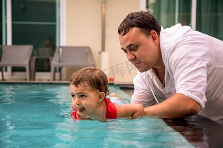 教游泳摄影照片_父亲教穿着泳装的可爱蹒跚学步的小女孩牵着他的手在游泳池里游泳。