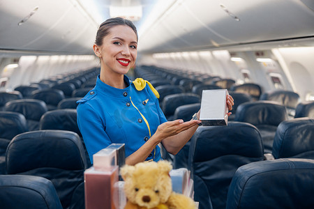 航空宣传摄影照片_身穿亮蓝色制服的漂亮空姐对着镜头微笑，一边宣传香水作为礼物，一边牵着手推车穿过空荡荡的飞机过道