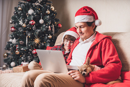 父亲带着孩子和小狗戴着圣诞老人的帽子，在圣诞节那天在笔记本电脑上进行视频通话，坐在客厅的沙发上，家里放着圣诞树。