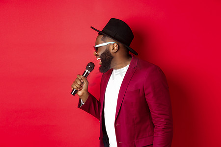 男唱歌摄影照片_热情的黑人男歌手在红色背景下表演，对着麦克风唱歌，穿着派对服装，站在红色背景上
