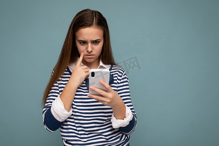 穿着休闲服的漂亮年轻女孩站在孤立的背景中，通过手机看手机屏幕在互联网上冲浪