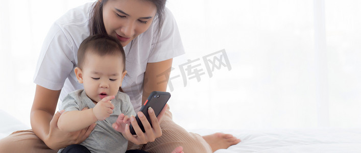电话视频摄影照片_年轻的亚洲母亲和小女婴或新生儿在卧室床上用智能手机自拍，幸福的妈妈和女儿在家里使用电话视频通话，两个人，家庭和沟通概念。