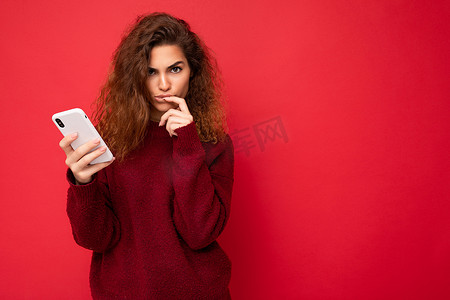 照片中，一头卷发的漂亮年轻女性穿着红色背景中突显的深红色毛衣，拿着手机看着相机，想着把手指放在嘴唇附近