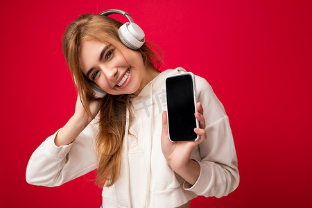 手机听音乐摄影照片_美丽快乐的微笑年轻女子穿着时尚休闲装，隔离在背景墙上，拿着手机，展示空显示屏，用于模型，戴着白色蓝牙耳机听音乐，开心地看着相机