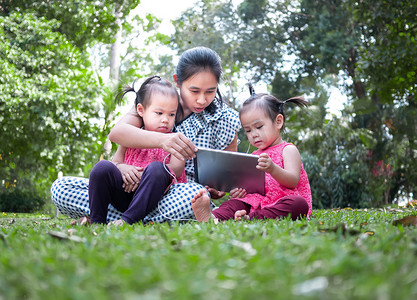 亚洲母亲和她的女儿们坐在花园的草地上，高兴地看着数字平板电脑。