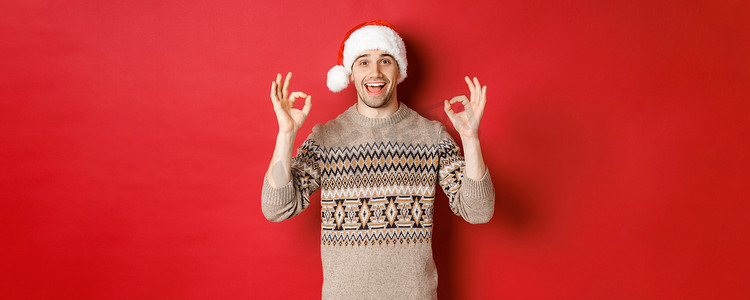 快乐而快乐的帅哥的画像，穿着冬衣和圣诞老人​​的帽子，表现出好的迹象，赞美圣诞晚会，站在红色背景上