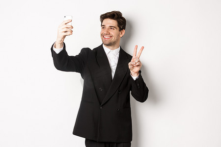 帅哥在新年派对上自拍、穿着西装、在智能手机上拍照并展示和平标志的肖像，站在白色背景下