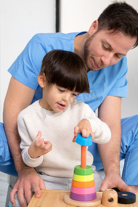 年轻的治疗师帮助患有脑瘫的可爱小男孩，在康复诊所玩开发玩具。