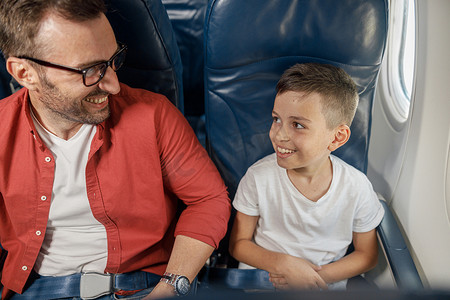 乘飞机旅行摄影照片_和父亲一起乘飞机旅行时，坐在窗边的微笑小男孩看起来很兴奋