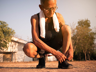 亚洲老人系着跑鞋的鞋带，准备在公园慢跑以保持健康。
