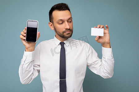 钱包银行卡摄影照片_英俊的男子穿着日常服装，隔离在背景墙上，手持并使用电话和信用卡付款，查看塑料银行卡