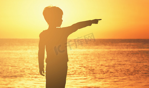 一个男孩在日出日落时在海洋橙色天空的海边的轮廓海洋男孩向侧面展示他的手指