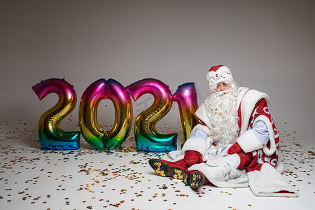 气球的2021摄影照片_圣诞老人坐在地上，拿着 2021 年形状的彩色气球、圣诞节摄影和新年广告