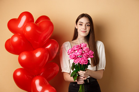 美丽的女孩拿着一束粉红玫瑰，对着镜头微笑，进行浪漫的约会，站在情人节心形气球旁边，米色背景