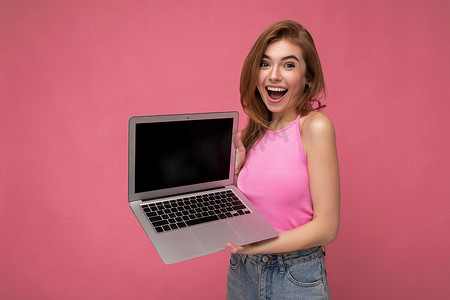 美丽的年轻金发女子拿着电脑笔记本电脑，身穿粉色短上衣，看着粉色背景中突显的相机