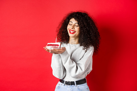 生日快乐庆祝蛋糕摄影照片_生日快乐的女孩庆祝和许愿，拿着生日蛋糕，微笑着，站在红色背景上