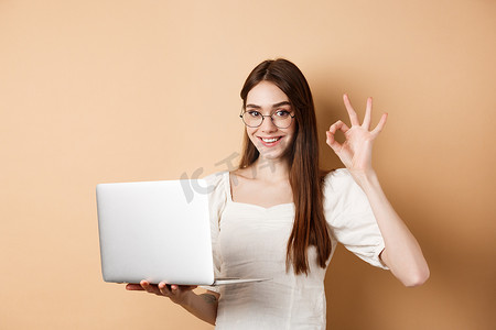 戴眼镜的自由职业女孩显示好的标志并在笔记本电脑上工作，保证质量好，在网上推荐一些东西，站在米色背景上