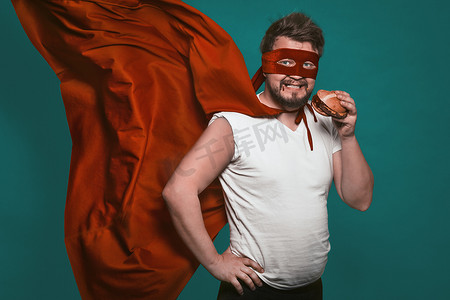 饥饿的超级英雄吃汉堡包，超级英雄或反英雄穿着红色西装，披着飞行斗篷，看着镜头吃快餐。
