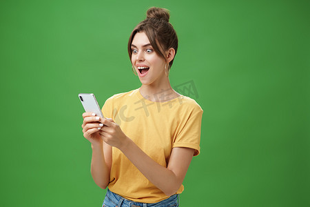 女人在智能手机上阅读令人惊讶的令人满意的信息，兴奋地张开嘴，微笑着惊讶地看着手机屏幕，在绿色背景下穿着休闲的黄色 T 恤