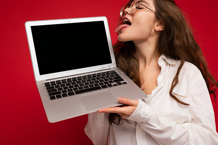 疯狂美丽的黑发卷发年轻女子手持电脑笔记本电脑，戴着眼镜，白色衬衫，舌头朝侧面看，红色背景上的舌头被隔离