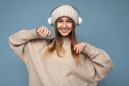 颈挂蓝牙耳机摄影照片_身穿米色冬季毛衣和帽子的迷人正面微笑的年轻金发女性与蓝色背景墙上隔开，戴着白色无线蓝牙耳机听音乐玩得开心，看着相机