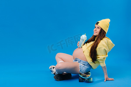 喝果汁的女孩摄影照片_蓝色背景的滑板上坐着一个穿着黄色衣服、喝着一杯果汁的怀孕女孩