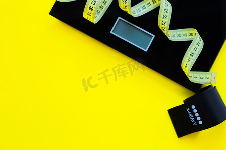 照片一侧的黄色背景上放着一个体重秤、一条黑色健身带和一条黄色卷尺。