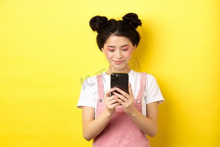 时尚的亚洲女孩，化妆迷人，用手机，对着屏幕微笑，站在黄色背景上
