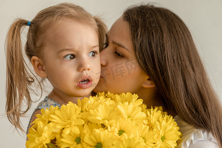母亲身份、母亲节、生日、童年、家庭观念-特写小女孩穿着裙子祝贺并给年轻的母亲一束黄色雏菊花，并在白色背景下亲吻她。