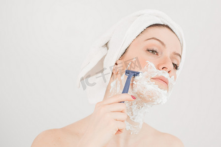 脸上的泡沫摄影照片_头戴毛巾、脸上剃须泡沫的快乐白人女性拿着白色背景上的剃刀