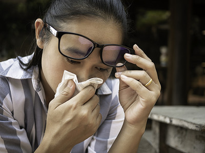 亚洲年轻女子拿着纸巾擦着眼镜，擤鼻涕。