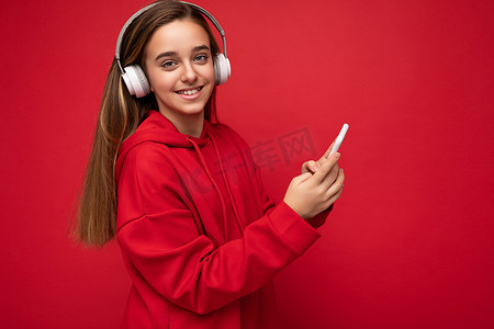 正面微笑的漂亮黑发女孩身穿红色连帽衫的侧面照片，在红色背景中被隔离，拿着并使用智能手机写短信，戴着白色无线耳机，听着很酷的音乐，看着相机