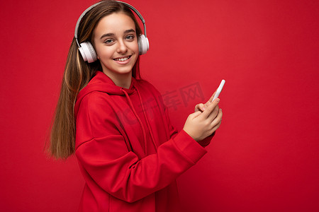 飞机侧面摄影照片_正面微笑的漂亮黑发女孩身穿红色连帽衫的侧面照片，在红色背景中被隔离，拿着并使用智能手机写短信，戴着白色无线耳机，听着很酷的音乐，看着相机