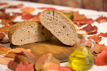 秋季乡村食品：在旧木切板上切成两半的整条面包、特级初榨橄榄油安瓿以及周围的红色和橙色紫藤叶