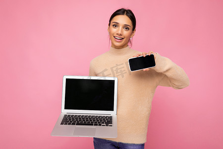 照片中，迷人漂亮、面带微笑、头发黑黑的年轻女性看着相机，拿着电脑笔记本电脑和手机，上面有空的监控屏幕，上面有模型和复制空间，穿着浅色毛衣，与粉色墙壁背景隔离