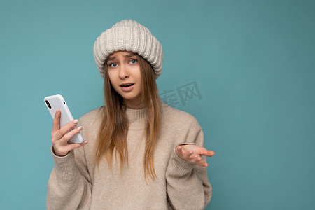 照片中，美丽的问心烦意乱的年轻金发女性身穿时尚的米色保暖毛衣，头戴蓝色背景中突显的冬季保暖米色针织帽，手拿空地，使用手机短信看着相机并提出问题