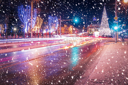 夜晚的城市有圣诞装饰、云杉和移动汽车前灯的痕迹，倒映在降雪的湿路上。