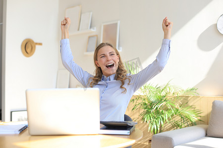 快乐的企业家女性坐在办公桌前阅读好消息，举起双手表达喜悦。