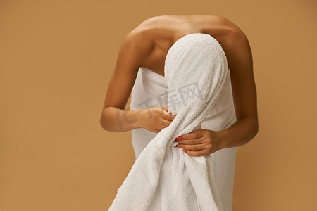 擦头发摄影照片_淋浴后的年轻女性站在米色背景中，用白毛巾擦头发