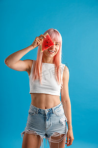 微笑的年轻女子，一头粉红色的头发，眼睛上拿着一片西瓜，站在蓝色的工作室背景中，与世隔绝