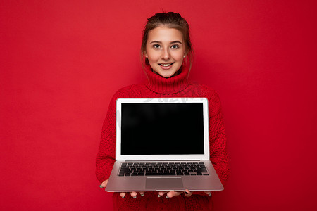 照片中，美丽而满意的快乐年轻女性拿着电脑笔记本电脑，看着穿着红墙背景中突显的红色毛衣的相机