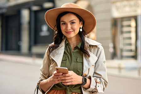 装秋摄影照片_一位年轻漂亮时尚女性的画像，身穿秋装，戴着帽子，手持智能手机，站在城市街道上看着相机微笑