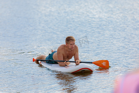 一名成年男子在躺着的冲浪板上游泳，用桨划水。
