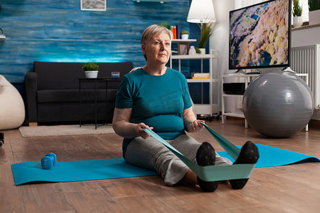 退休老年妇女坐在瑜伽垫上用弹力带拉伸腿部肌肉