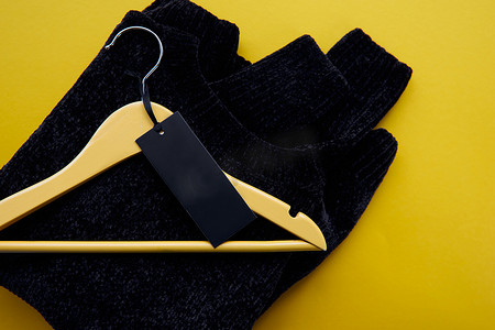 服装海报背景摄影照片_木制衣架与黑色舒适套头衫和纸标签隔离在黄色纸背景。