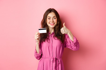 黑发女孩竖起大拇指微笑，推荐银行，拿着塑料信用卡，站在粉色背景下